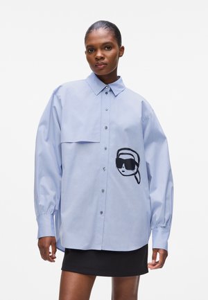 Light blue button-up shirt with a left patch pocket, featuring a black illustrated face design on the front. Long sleeves with gathered cuffs.