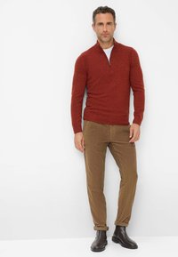 Roter gestrickter Pullover mit einem Halbreißverschluss-Kragen, kombiniert mit braunen Cordhosen und schwarzen Knöchelstiefeln. Glatte Textur mit feinen Sprenklern.