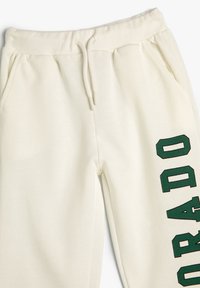 Spodnie dresowe w kolorze ecru z elastycznym pasem, sznurkiem ściągającym, kieszeniami bocznymi oraz zielonym pionowym napisem "ORADO" na prawej nogawce.