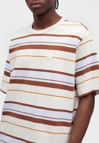 Homme portant un T-shirt blanc avec des rayures horizontales marron, blanches et orange et un petit logo vert sur la poitrine.