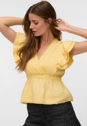 Femme aux longs cheveux bruns portant un chemisier jaune à manches courtes volantées et un pantalon foncé, posant avec les bras levés.