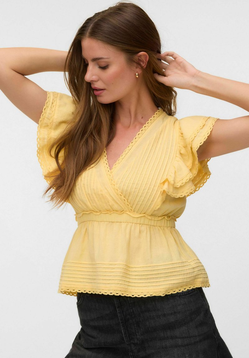 Femme aux longs cheveux bruns portant un chemisier jaune à manches courtes volantées et un pantalon foncé, posant avec les bras levés.