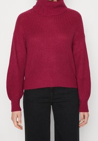 Nainen, joka käyttää tekstuurilla varustettua pitkähihasta viininpunaisesta turtleneck-puserosta mustien housujen kanssa, seisoo yksinkertaista taustaa vasten.