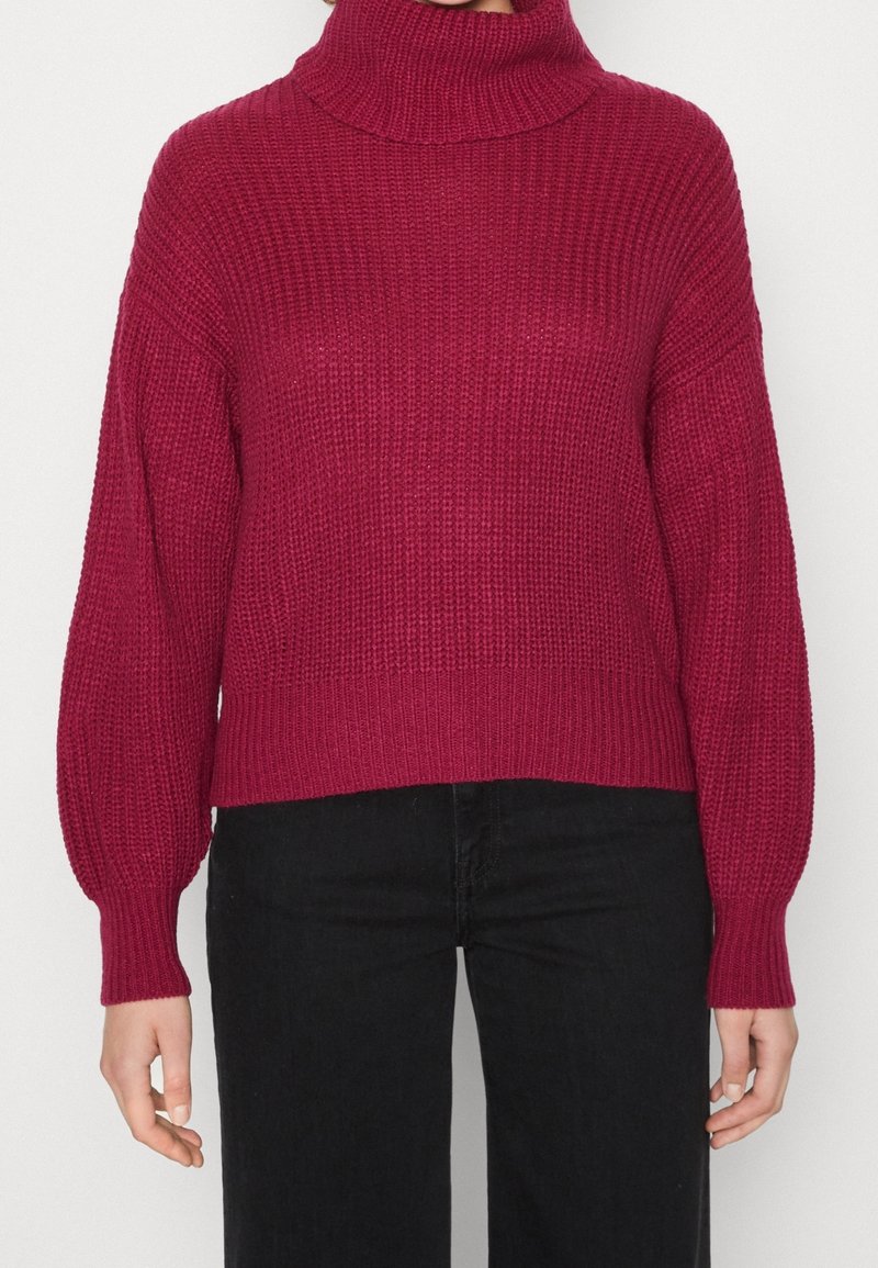 Nainen, joka käyttää tekstuurilla varustettua pitkähihasta viininpunaisesta turtleneck-puserosta mustien housujen kanssa, seisoo yksinkertaista taustaa vasten.