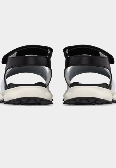 Ζευγάρι ασπρόμαυρων αθλητικών σανδαλιών με ρυθμιζόμενους ιμάντες και χοντρές υφασμάτινες σόλες, εμφανιζόμενα από πίσω σε λευκό φόντο.