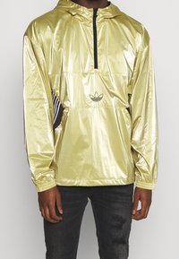 Hombre vestido con una chaqueta cortaviento brillante de color dorado claro con una cremallera negra y el logo de Adidas, combinada con jeans negros desgastados.
