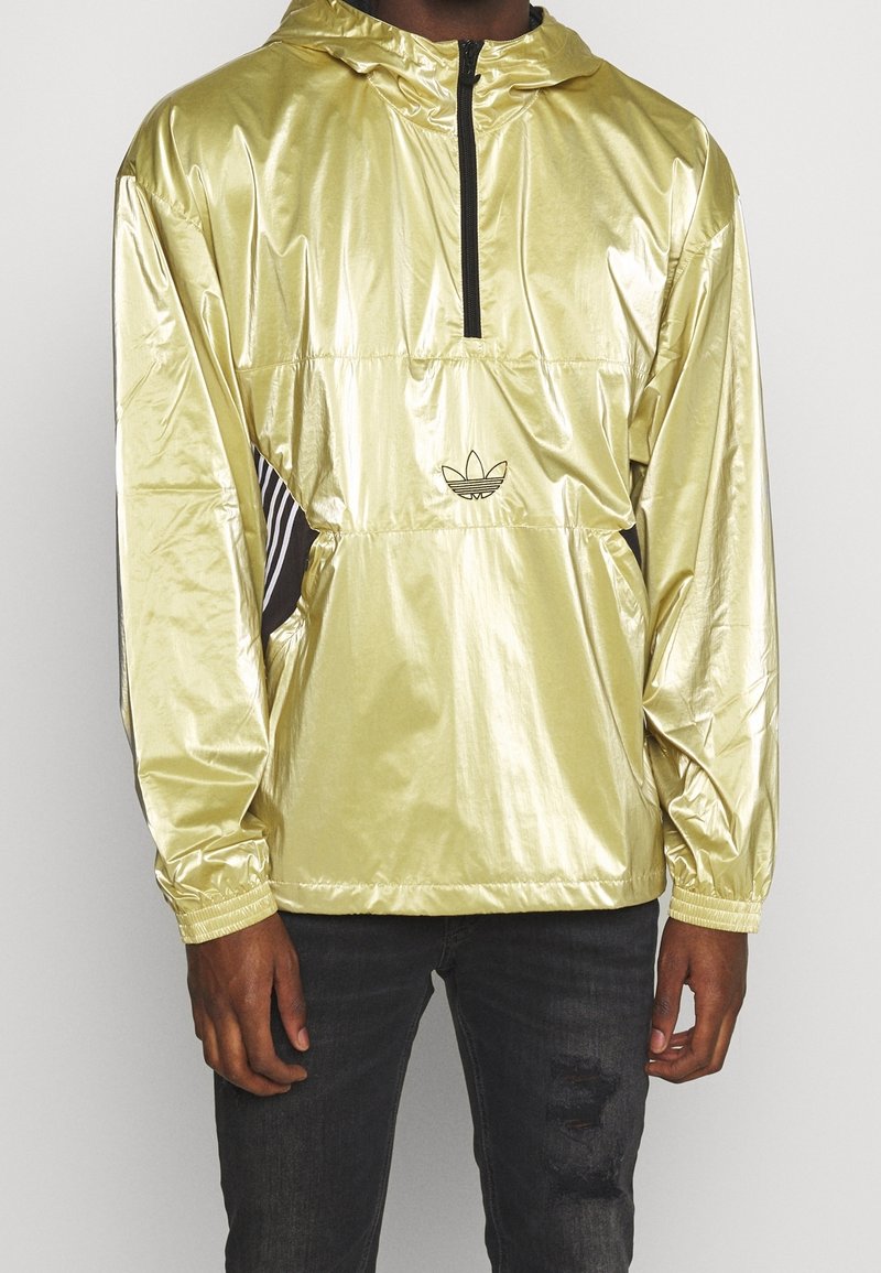 Hombre vestido con una chaqueta cortaviento brillante de color dorado claro con una cremallera negra y el logo de Adidas, combinada con jeans negros desgastados.