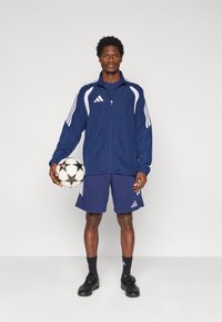 Μπλε σκούρο αθλητικό μπουφάν με λευκές λεπτομέρειες, ταιριαστά σορτς και μαύρα ποδοσφαιρικά παπούτσια. Κρατώντας μια ποδοσφαιρική μπάλα με σχέδιο.