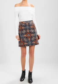Femme portant un haut blanc à manches longues dénudées, une jupe à motif floral bleu marine et des bottines noires, se tenant devant un fond uni.
