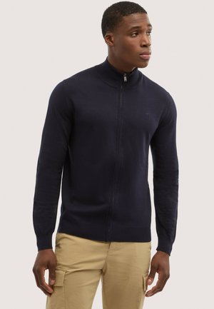 Uomo che indossa un maglione con zip color blu navy e pantaloni cargo beige, in piedi e che guarda di lato su uno sfondo semplice.
