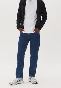 Černý cardigan zapnutý přes bílé tričko, zkombinovaný s modrými dž íny a stříbrnými sportovními botami s modrými detaily.