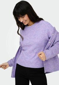 Pull violet en maille avec un col rond, tissu texturé et détails de boutons sur des manches ouvertes, associé à un jean noir.