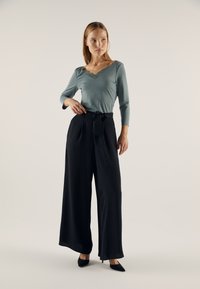 Light blue long-sleeve top with lace trim and V-neck, paired with high-waisted black wide-leg trousers secured with a tie belt. Black pointed shoes.