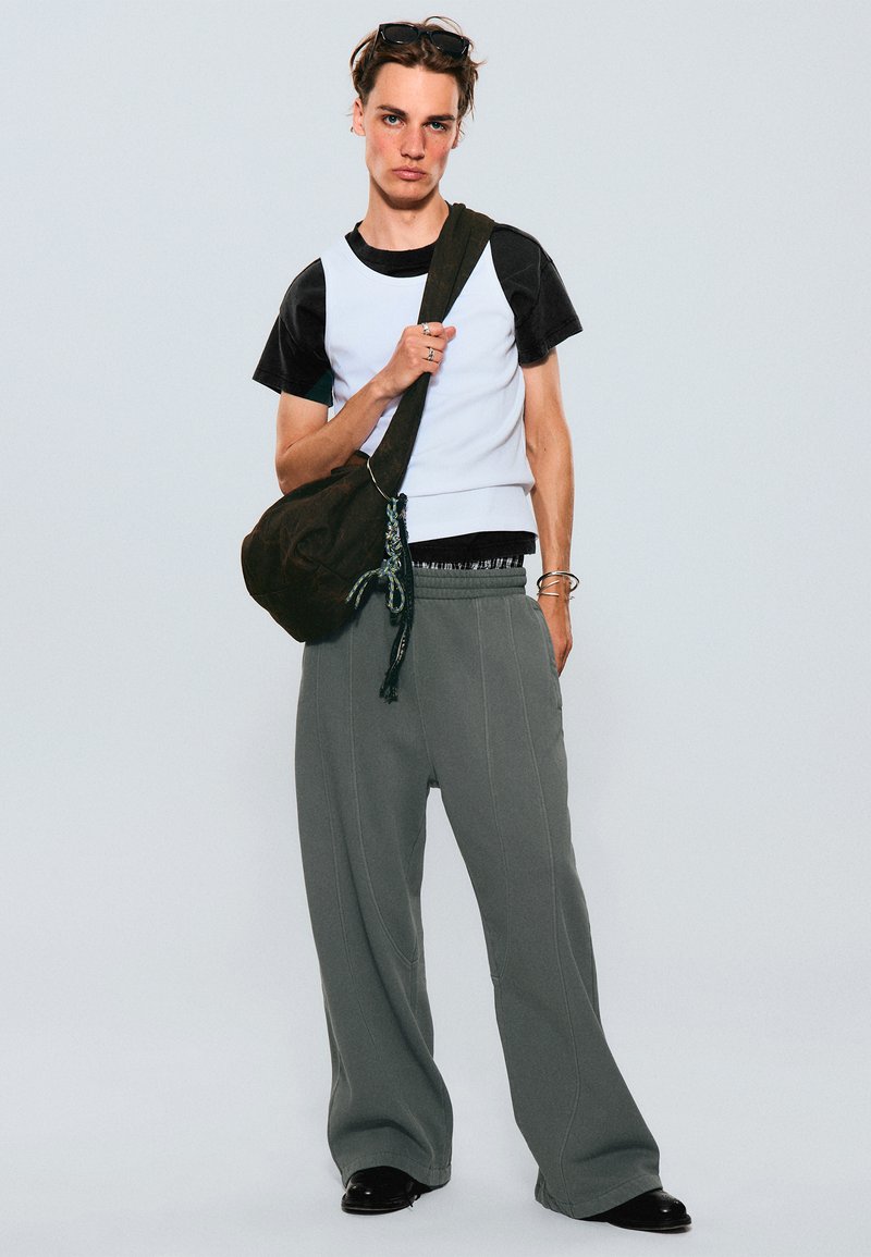 Chemise à manches courtes blanche et noire superposée à un pantalon de survêtement gris ample. Accompagnée d'un sac à épaule marron foncé avec des accents décoratifs.