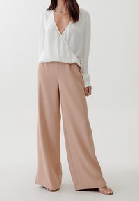 Vit, långärmad, insvängd topp i kombination med vida, beige byxor. Mjuk tyg, slät textur och en djup V-ringning.