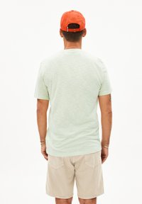 Hellegrünes Kurzarmhemd mit strukturiertem Finish, kombiniert mit beigen Shorts. Die Person trägt eine orangefarbene Kappe, die von hinten sichtbar ist.