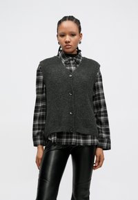 Γκρι πλεκτό γιλέκο με V-λαιμόκοψη, πέντε κουμπιά και ριμπ τελείωμα, φορεμένο πάνω από πουκάμισο με ασπρόμαυρο καρό σχέδιο. Συνδυασμένο με μαύρο δερμάτινο παντελόνι.