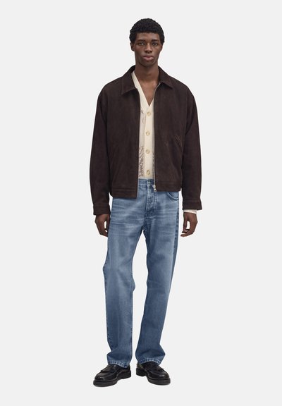 Homme debout portant une veste zippée marron foncé, un cardigan crème boutonné, un jean bleu clair et des chaussures noires sur un fond blanc.