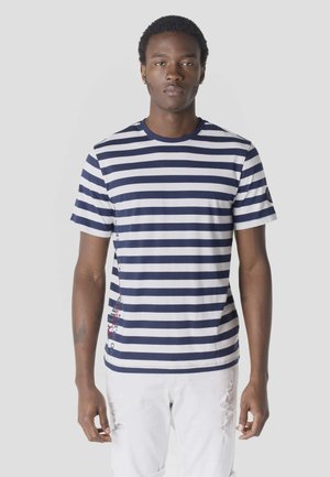 Jonge man die een marineblauw-wit gestreept t-shirt met horizontale strepen en witte versleten spijkerbroek draagt, staand tegen een effen lichte achtergrond.