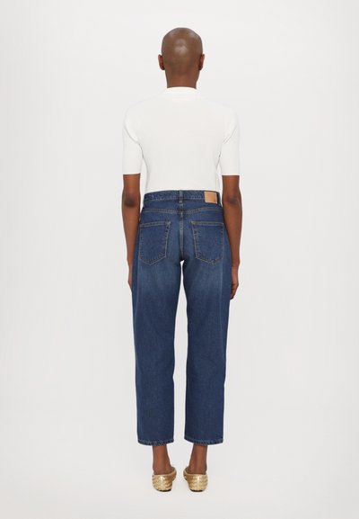 Jeans de denim azul de cintura alta con pierna recta, detalles de costura sutiles y bolsillos traseros. Combinados con una camisa blanca ajustada y zapatos dorados.