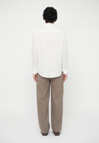 Mann med krøllete hår iført en hvit langermet skjorte, løse brune bukser og brune sko, stående med ryggen mot en ensfarget bakgrunn.