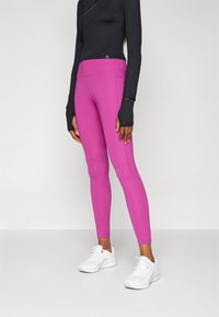 Fitting fekete hosszú ujjas felső, hüvelykujjlyukakkal, élénk rózsaszín leggings-szel párosítva. Az edzőcipők fehérek, texturált felülettel és fűzővel.