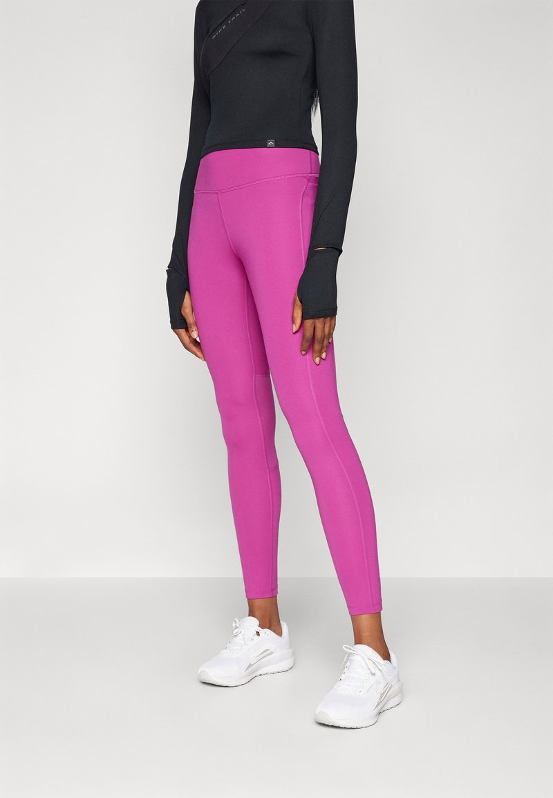 Fitting fekete hosszú ujjas felső, hüvelykujjlyukakkal, élénk rózsaszín leggings-szel párosítva. Az edzőcipők fehérek, texturált felülettel és fűzővel.