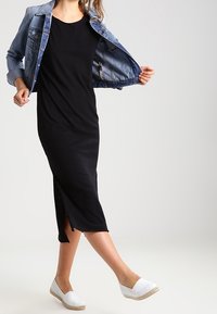 Svart midiklänning med rund halsringning och sidoklutningar, i kombination med en denimjacka och vita espadrilloskor. Slät yta, avslappnad design.