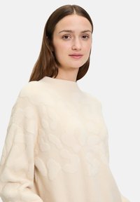 Cremfarbener Pullover mit strukturiertem, geprägtem Leopardenmuster. Verfügt über einen hohen Kragen und überschnittene Schultern. Weicher, plüschiger Stoff.