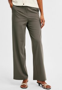 Vida leggings i olivgrön tyg med slät textur. Sidofickor och en främre veckdetalj. Matcher med svarta sandaler med remmar.