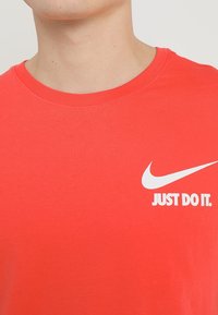 Osoba v jasně červeném tričku s kulatým výstřihem, na kterém je bílé logo Nike a text "JUST DO IT." na hrudi.