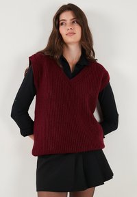 Burgundzki dzianinowy sweter bez rękawów z dekoltem w serek, z prążkowaną teksturą, noszony na czarnej koszuli z kołnierzykiem i czarnej spódnicy.