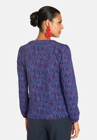 Langärmlige Bluse aus dunkelblauem Stoff mit einem floralen und geometrischen Muster in Rot und Weiß; verfügt über einen runden Ausschnitt und geraffte Ärmel.