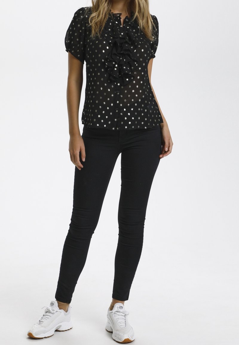 Femme portant un chemisier noir à pois avec des volants, un pantalon noir cintré et des baskets blanches, debout sur fond blanc.