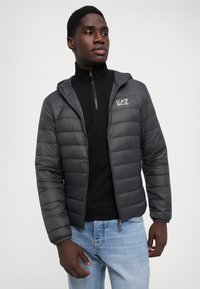 Doudoune noire avec capuche, dotée d'une fermeture éclair à l'avant et du logo EA7 sur la poitrine, associée à un haut noir à col montant et un jean en denim clair.