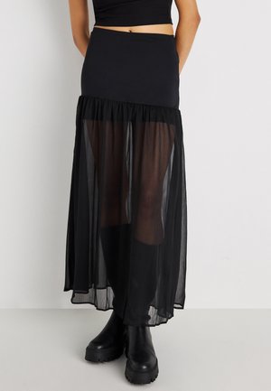 Persona que lleva una falda negra de cintura alta con una capa transparente inferior y botas negras gruesas hasta el tobillo, de pie frente a un fondo liso.