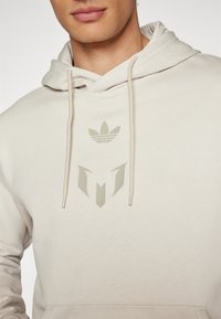 Beige Kapuzenpullover mit Kordelzug, aus strukturiertem Stoff und mit zwei Logos – eines über der Brust und eines in der Mitte, beide in einem gedeckten Farbton.