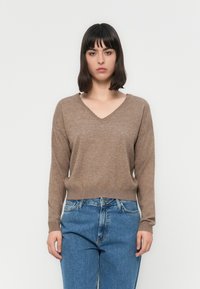 Donna con capelli castani a spalla che indossa un maglione marrone a V e jeans blu su uno sfondo bianco semplice.