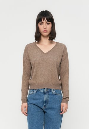 Frau mit schulterlangem dunklem Haar, die einen braunen V-Ausschnitt-Pullover und blaue Jeans vor einem schlichten weißen Hintergrund trägt.