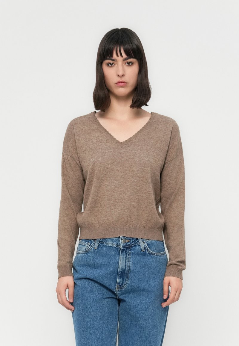 Donna con capelli castani a spalla che indossa un maglione marrone a V e jeans blu su uno sfondo bianco semplice.