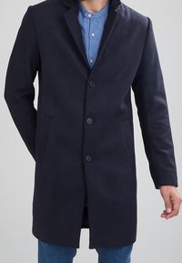 Cappotto di lana blu navy con collo a risvolto, chiusura monopetto, due tasche laterali e texture liscia, abbinato a una camicia di jeans sotto.