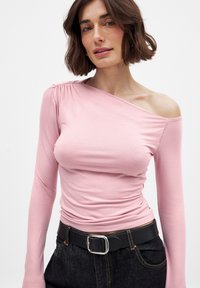 Roze langiedig top met een schouder bloot, met geplooide details bij de schouder. Draagt een zwarte riem over donkere denim.