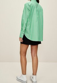 Osoba ubrana w zieloną i białą pionową paskową koszulę z długim rękawem, czarną dżinsową mini spódniczkę oraz białe trampki za kostkę, stoi tyłem.