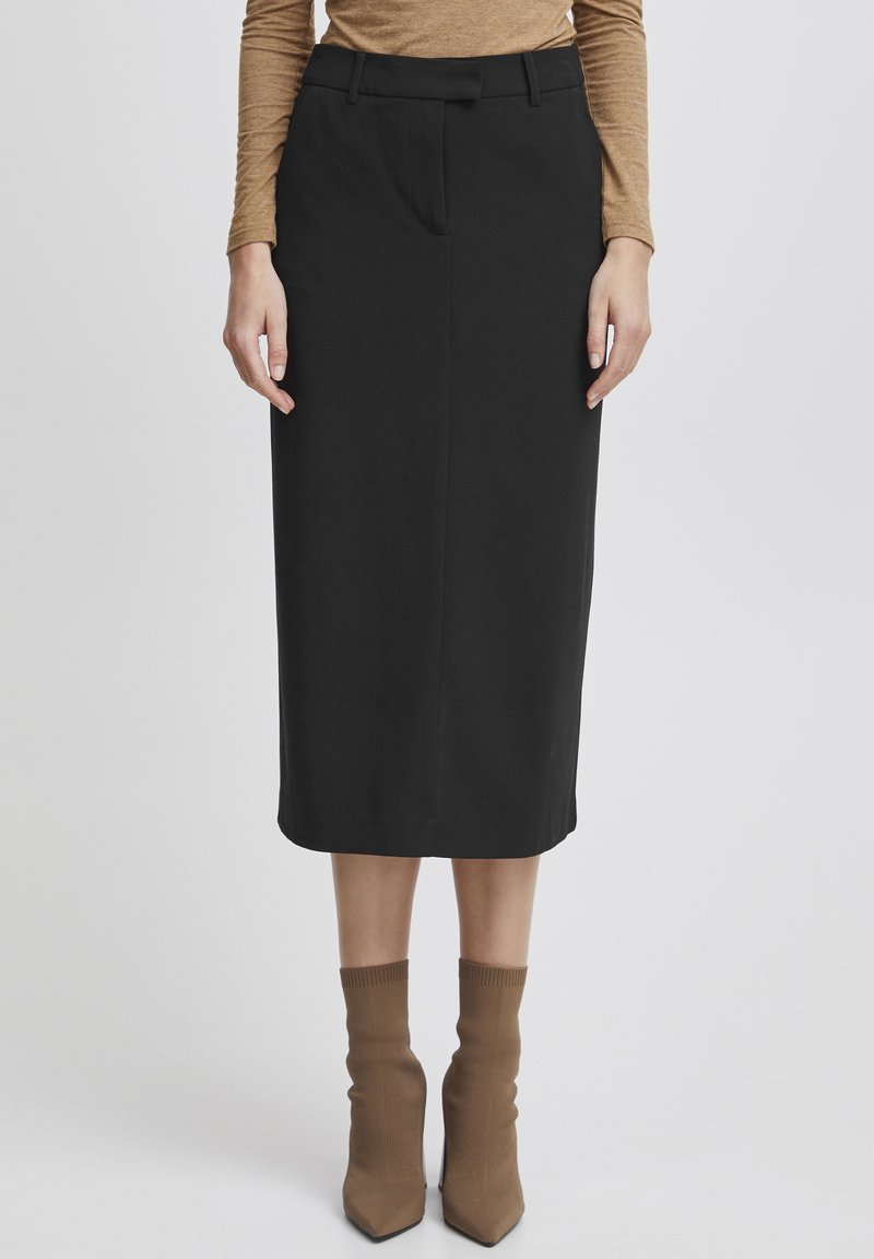 Jupe midi noire avec une coupe droite, des plis à l'avant et des passants de ceinture. Associée à des chaussettes en maille marron jusqu'à la cheville et des talons aiguilles.