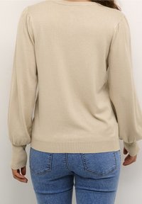 Béžový pulovr s nařasenými detaily na rukávech a žebrovanými manžetami, doplněný modrými džínami s kapsami vzadu a přiléhavým střihem.