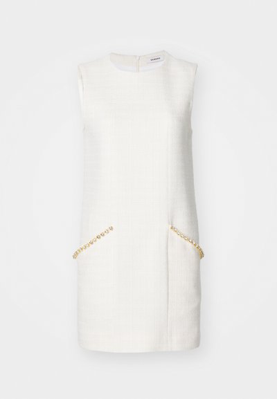 Valkoinen hihaton mekko, jossa on teksturoitu kangas, ja jossa on kaksi etutaskua, jotka on koristeltu kultaketjuilla, sekä takana vetoketjusuljenta.