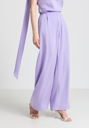 Donna che indossa pantaloni larghi color lavanda e top coordinato con una lunga fascia, abbinati a sandali bianchi con tacco alto e punta aperta.