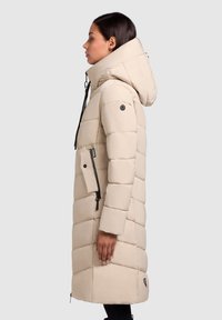 Vrouw in een lange beige pufferjas met een grote capuchon, met een ritszak en zijventilatie, staande in profiel tegen een lichte achtergrond.