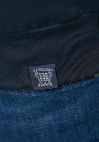 Marineblaues Kleidungsstück mit geripptem Kragen; verfügt über ein aufgenähtes Etikett, das "U.S. Polo Assn. 135 Jahre" mit dekorativen Elementen zeigt.