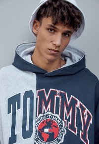 Hettegenser med grå og marine paneler, en stor "TOMMY" trykk, globusgrafikk og teksturert stoff. Brukt med en hvit bøttehatt.